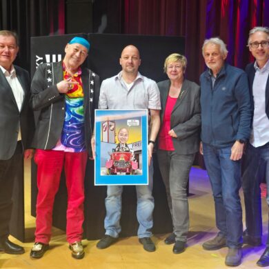 Auf der Bühne im Festsaal: Gruppenfoto mit dem Bürgermeister und dem Karrikaturisten. In der Mitte zeigt Walter Wistermayer eine Karrikatur von sich selbst;